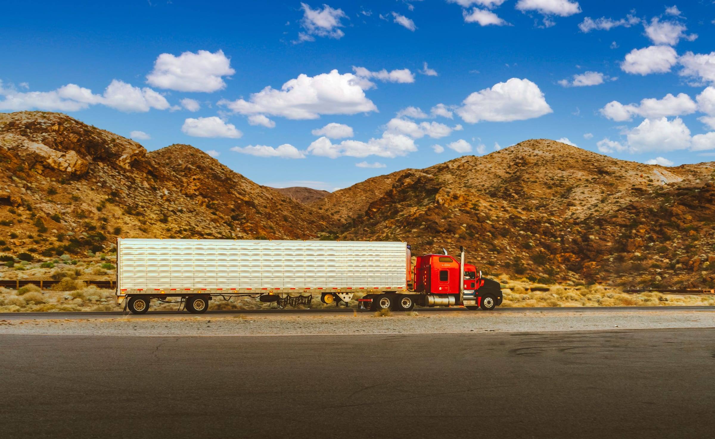 Tractocamión rojo con un remolque blanco largo circula por una carretera del desierto en Nevada, con montañas rocosas y cielo azul con nubes.